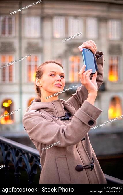 Elegant, young woman taking a photo with her cell phone camera while traveling (shallow DOF; color toned image)