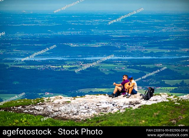 16 August 2023, Bavaria, Aschau Im Chiemgau: A hiking couple sits on a rock at the Kampenwand and enjoys the view into the valley and the nearby Chiemsee