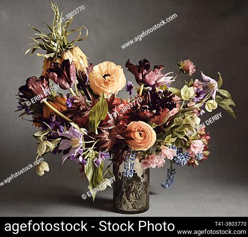 A bouquet of red, orange, purple, green and blue flowers in a mercury glass vase against a grey backdrop