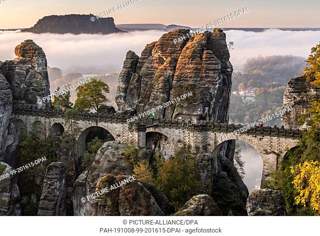 07 October 2019, Saxony, Rathen: In the morning fog lies over the Saxon Switzerland National Park behind the bastion bridge built in 1851 and the 415-metre-high...