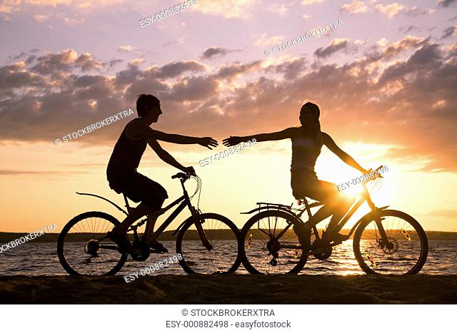 Silhouettes of happy couple stretching arms to each other while riding bicycles on seashore at sunset