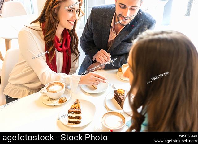 High-angle view of a happy couple sharing cakes while sitting at table with their mutual friend in a trendy coffee shop