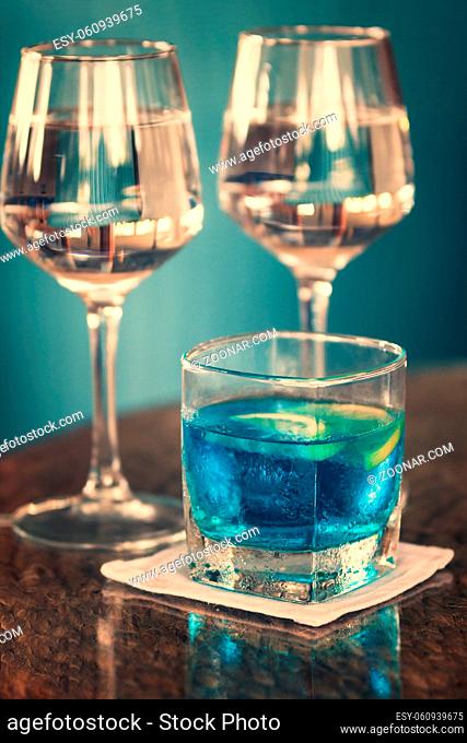 Longdrink mit Blue Curacao und zwei Gläser mit Roséwein auf glänzendem Holztisch vor türkisfarbener Wand