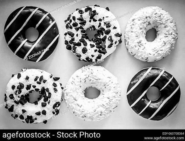 Flat lay image of six ring donuts with white glaze and hundreds and thousands, chocolate and stripes and white glaze with black cookies, black and white image