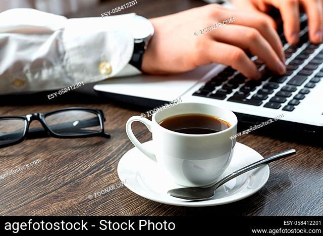 Businessman sitting at desk and working with laptop computer. Close-up of man hands typing on keyboard in office. Businessman workplace with glasses and cup of...