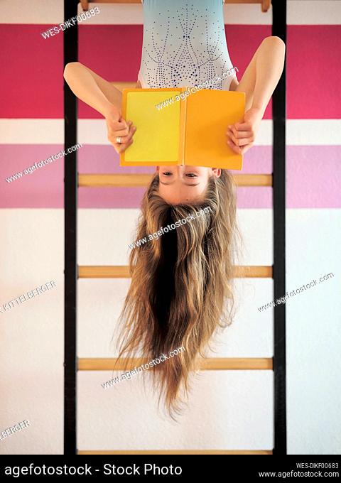 Girl reading book hanging upside down on wall bars