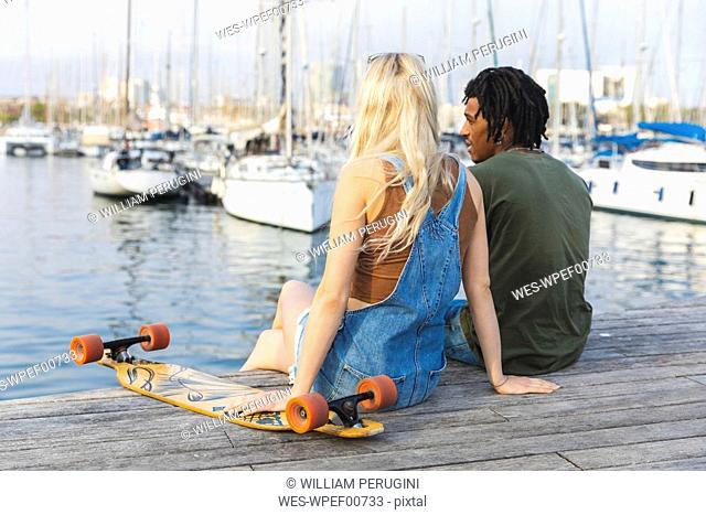 Spain, Barcelona, multicultural young couple with longboard relaxing at harbour
