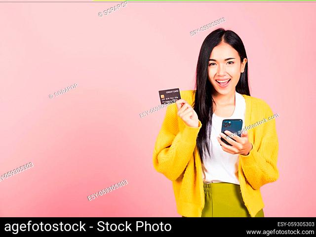 Happy Asian portrait beautiful cute young woman excited smiling hold mobile phone and plastic debit credit bank card, studio shot isolated on pink background