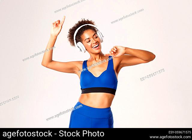 Sport and lifestyle concept. Happy attractive african-american fitness woman dancing, listening music in headphones during workout, white background