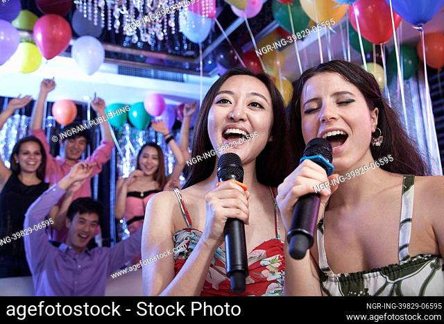 Two friends holding microphones and singing together at karaoke, friends in the background