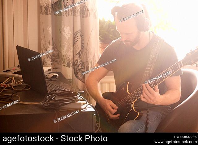 Bearded man musician playing music and composing a song with electric guitar piano and laptop computer while sitting in living room