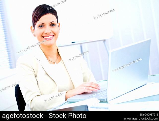 Businesswoman working on laptop computer in brightly lit office, smiling, looking at camera
