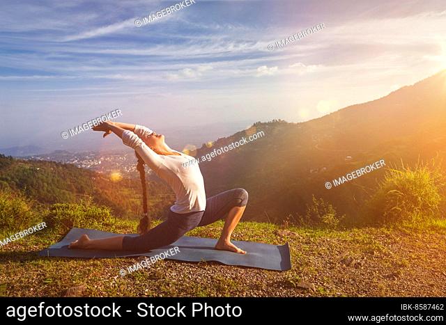 Yoga outdoors, sporty fit woman practices yoga Anjaneyasana, low crescent lunge pose outdoors in mountains in morning. With light leak and lens flare