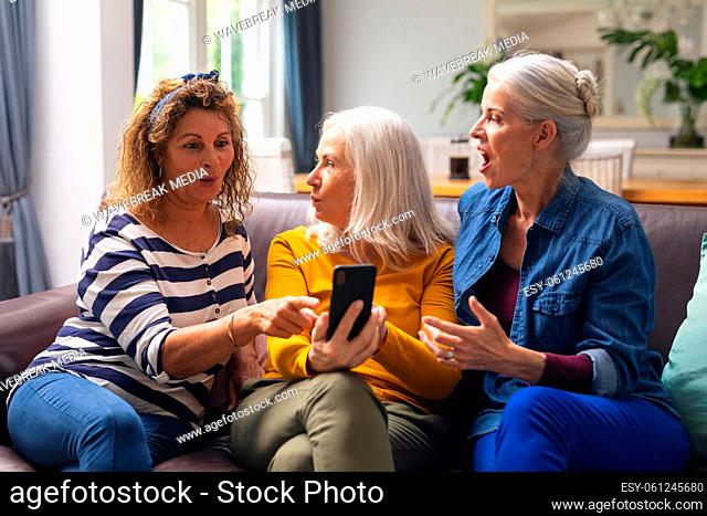 Surprised multiracial senior female friends looking at smart phone while sitting on sofa at home