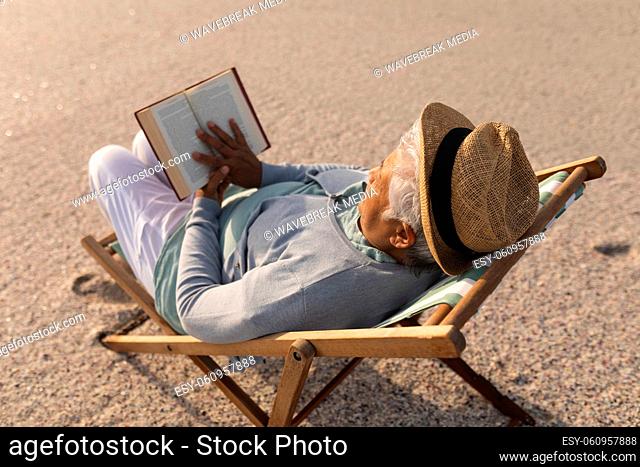 Biracial senior woman wearing hat reading book while relaxing on folding chair at beach