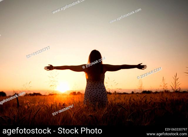 Back view woman with long hair in field at sunset, spreading out her hands, greeting sun. Young girl living in contact with nature