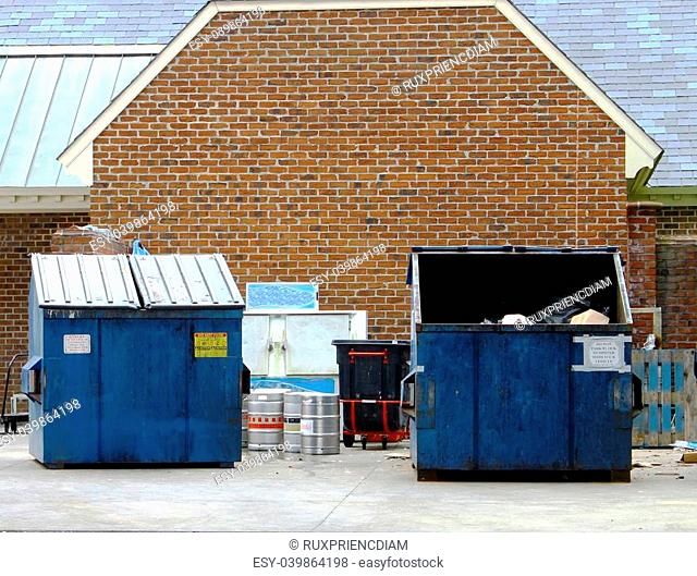 A couple of large blue trash dumpsters with a trash can, some Kegs and an Ice locker behind a business