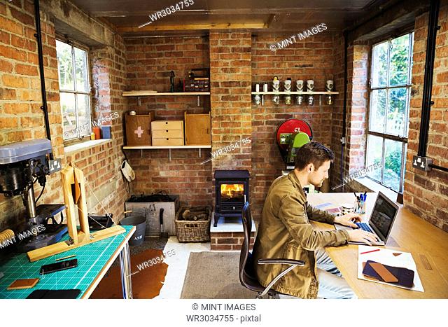 A designer seated in his leatherwork workshop, at a desk using a laptop. Woodburning stove with a glowing fire lit
