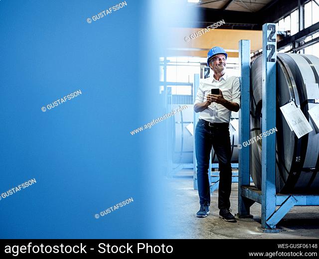 Smiling male engineer with mobile phone standing by rolled up metal sheet