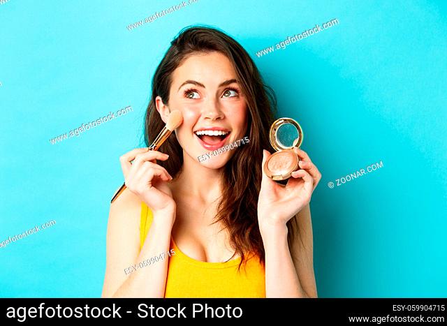 Beauty. Close up of beautiful young woman look upper left corner and smile while applying blushes on cheeks with make up brush