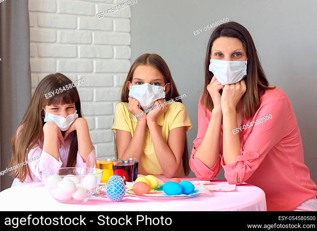 Mom and two daughters at the table in medical masks paint Easter eggs for the holiday