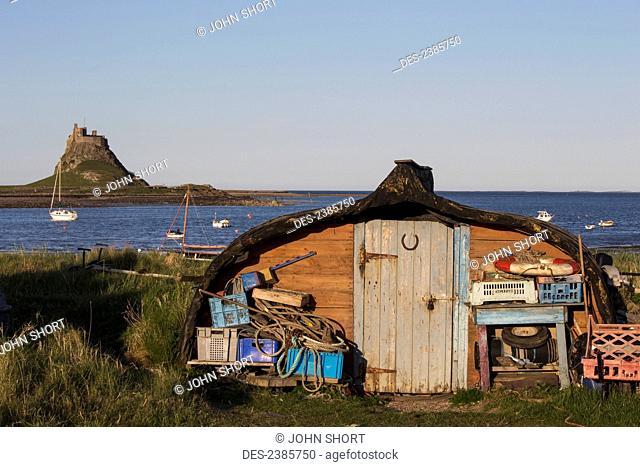 A shed with fishing equipment at the water's edge with a view of Lindisfarne Castle on Holy Island; Lindisfarne, Northumberland, England