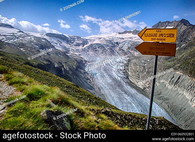 mountain landscape with green fields and snowy peaks and wild glacier in the Swiss Alps and yellow hiking trail signpost in the Zinal Valley in the Valais in...