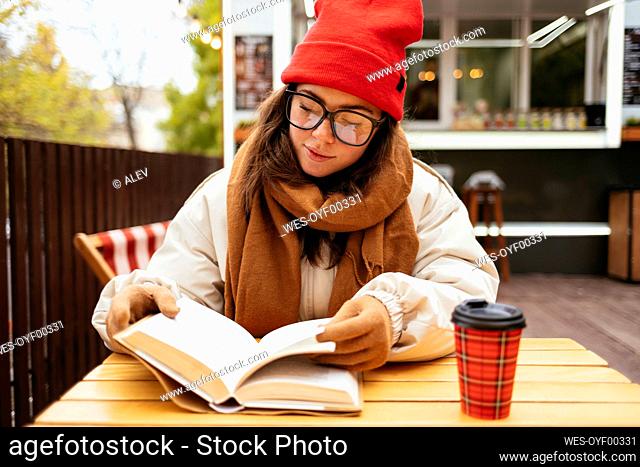 Woman wearing eyeglasses reading book while sitting at sidewalk cafe