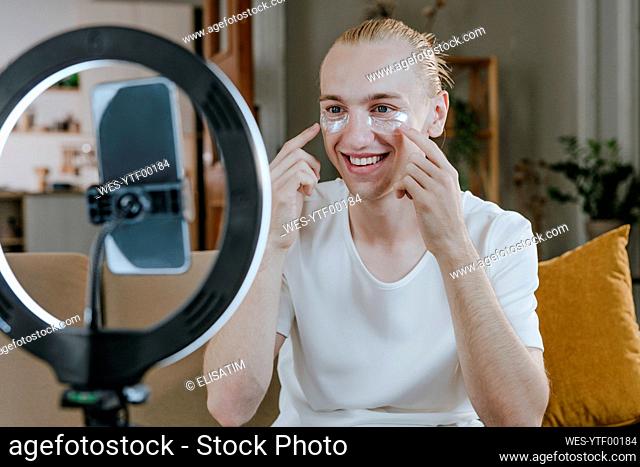 Happy young influencer applying medical eye patch and vlogging through mobile phone at home