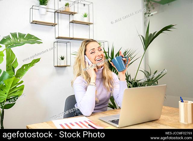 Cheerful young female professional talking on smart phone while drinking coffee in office