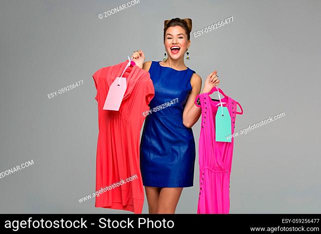 Beautiful happy woman holding two dresses on hangers. Price tags. Over grey background. Copy space