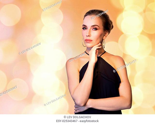 people, luxury, jewelry and fashion concept - beautiful woman in black wearing diamond earrings over beige holidays lights background
