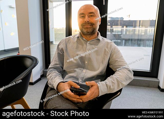 Portrait of mature businessman sitting on chair in office with mobile phone