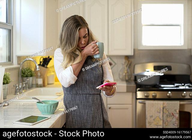 Coughing young woman in her kitchen eating soup and drinking tea, checking smartphone