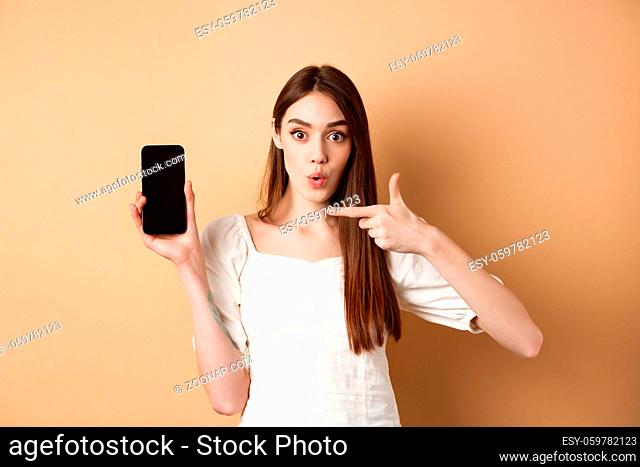 Excited woman showing news on screen, pointing at empty phone and looking surprised, standing on beige background