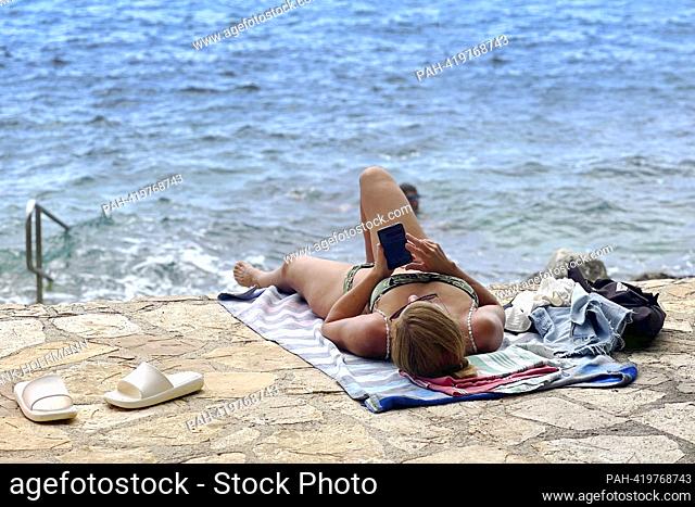 Plava Laguna, Adriatic coast near Porec / Croatia, beach. Vacationer, young woman, single looks at her smartphone, cell phone, sunbathes on the stone beach