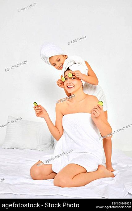 smiling woman with daughter in white towels holding fresh cucumber slices
