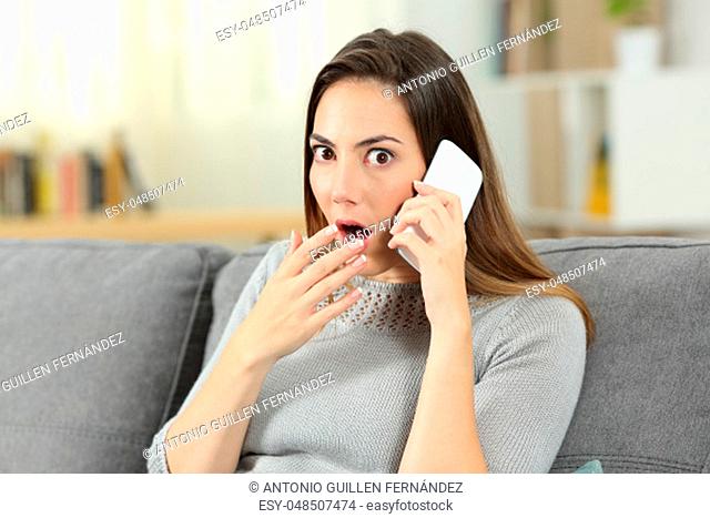 Amazed woman talking on the phone looking at you sitting on a couch in the living room at home