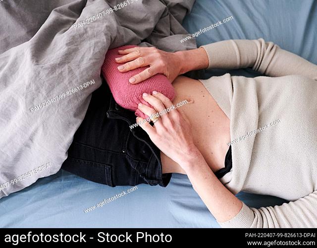 PRODUCTION - 30 March 2022, Berlin: ILLUSTRATION - A woman holds a hot water bottle to her lower abdomen while lying in bed