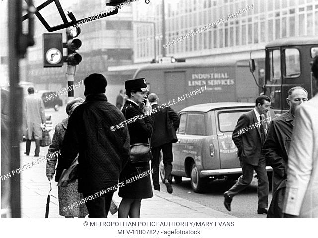 Woman police officer (Barbara Gelder) speaking into a radio on a busy London street full of pedestrians and traffic. She is wearing the new style Norman...
