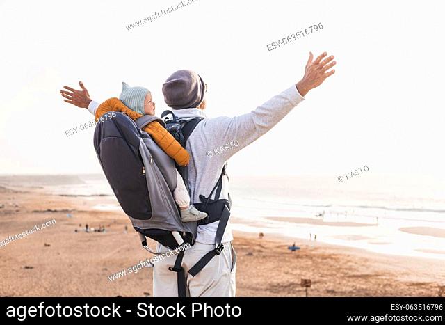 Young father rising hands to the sky while enjoying pure nature carrying his infant baby boy son in backpack on windy sandy beach