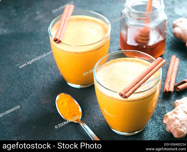 Healthy drink golden turmeric latte in glass.Gold milk with turmeric, ginger root, cinnamon sticks, turmeric powder and honey over black cement background