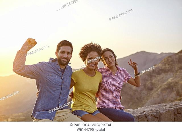 Three celebrating friends posing for a picture in the late afternoon sunshine on a mountain them while wearing casual clothing
