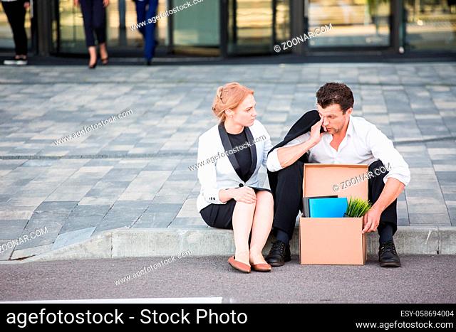 Fired business man sitting frustrated and upset on the street near office building with box of his belongings. He lost work