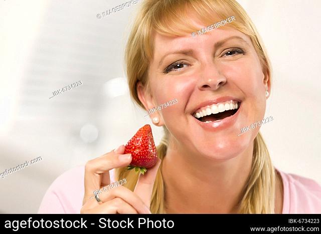 Pretty smiling blonde woman holding strawberry in her kitchen