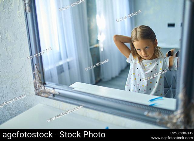 Pretty Little girl combing by herself hair in front of a mirror, brushing her hair at home, child smiling, morning routine