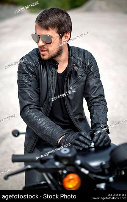 Handsome man with a beard on the black motorcycle on the blurry background outdoors. His hands lies on the motorcycle. He looks to the left