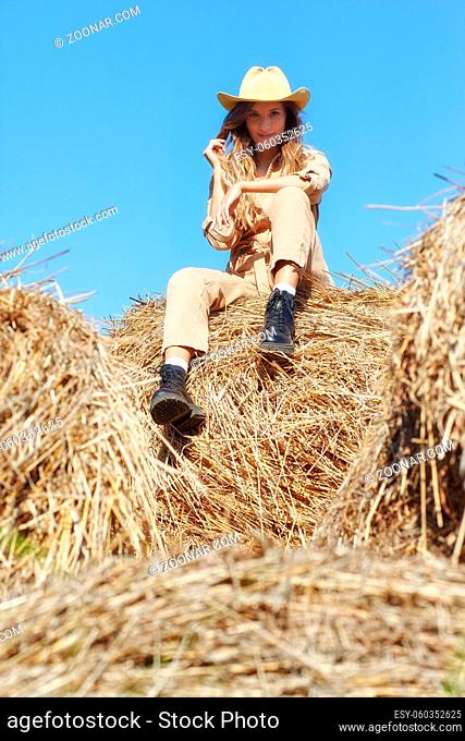 Young blonde woman in a beige jumpsuit is sitting on the rolls of hay. Romantic country girl in cowboy hat on hay