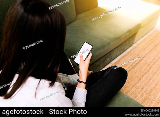 Communication concept with black hear girl sitting on green sofa and holding smartphone in her hand with empty white screen, sun beam on the front