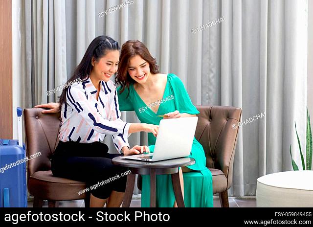 Young woman with luggage pointing at laptop computer screen, show her friend the product they looking for on internet. Caucasian woman holding credit card...